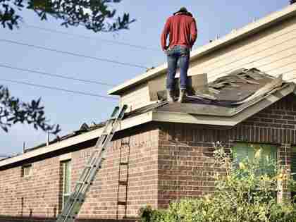 A person standing on a partly stripped residential roof beside a ladder.